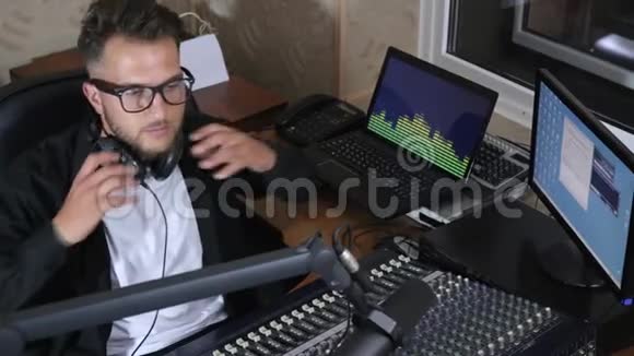 戴着耳机的DJ坐在混合控制台旁边在背景电脑屏幕上对着迈克说视频的预览图