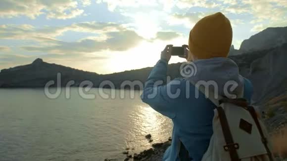 年轻迷人的女人从山顶看日落或从海湾升起女孩拿起全景照片打电话视频的预览图