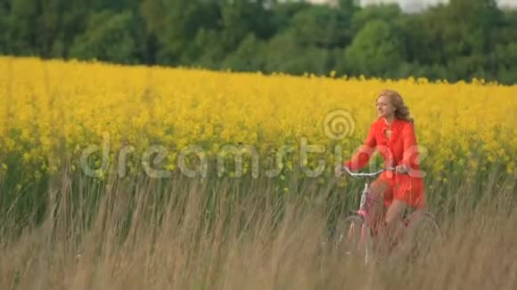 一位穿着红裙子的漂亮金发女郎正快乐地骑着自行车沿着盛开的黄色田野视频的预览图