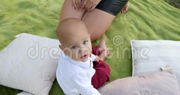 穿着西装的婴儿漂亮的大眼睛看着坐在公园户外枕头附近的绿色格子上的相机视频的预览图