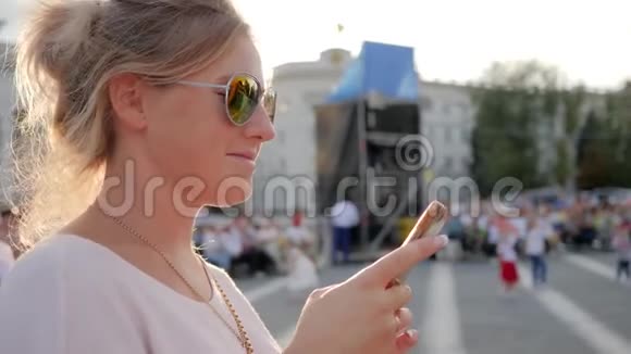 戴着眼镜戴着智能手机的金发眼镜的年轻女子在街上牵手视频的预览图