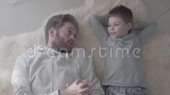 大胡子的父亲和他的小儿子躺在白色的毛皮地毯上进行有趣的谈话父亲子女视频的预览图