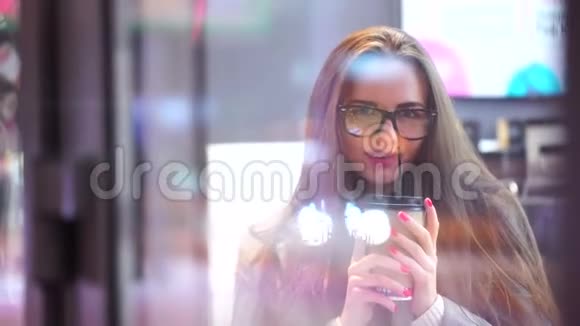 戴眼镜的快乐女孩喝着咖啡或茶在咖啡馆里放松微笑着透过窗户咖啡馆看着相机视频的预览图
