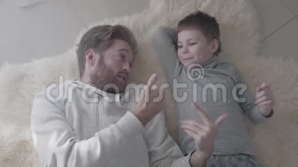 大胡子的爸爸和他的小儿子躺在白色的毛皮地毯上进行了有趣的交谈父子视频的预览图
