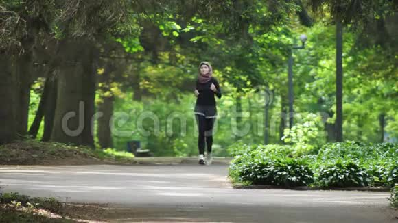 戴着头巾跑步的年轻运动女孩在绿色公园慢跑前视图50fps视频的预览图