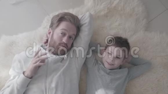 有胡子的父亲和他的小儿子躺在白色的毛皮地毯上进行有趣的谈话父亲子女视频的预览图