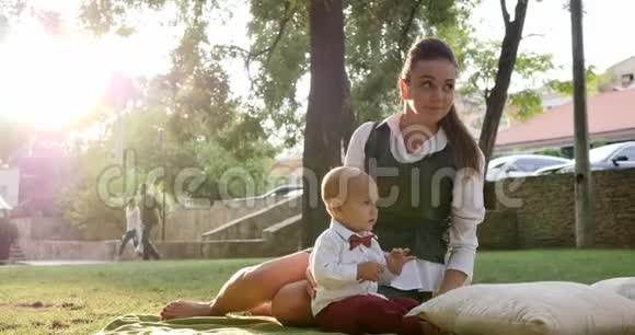 年轻的母亲坐在公园的绿色草坪上带着可爱的西装革履的婴儿和蝴蝶结视频的预览图