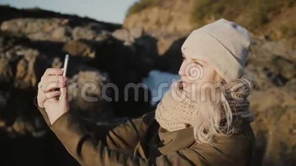 年轻的金发女人站在山上在美丽的背景下在智能手机上自拍视频的预览图