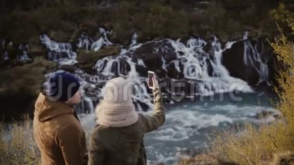 年轻的旅行夫妇站在瀑布附近的山谷在智能手机上聊天或自拍照片视频的预览图