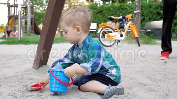 可爱的2岁幼儿男孩在公园里挖沙子和倒在桶里的慢动作视频视频的预览图