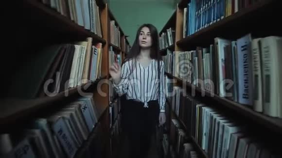 漂亮的小女孩留着长长的黑发在图书馆的书架之间慢动作视频的预览图