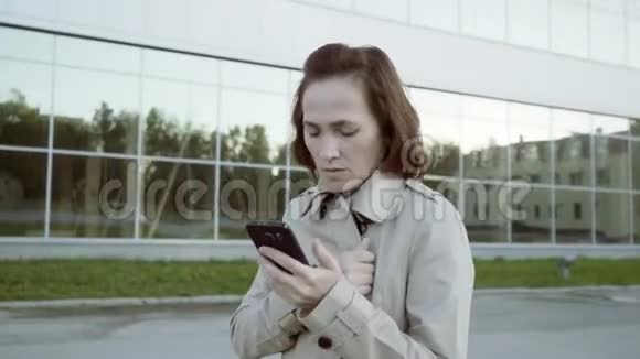 一位身着米色战壕的女商人手里拿着智能手机的肖像经理拿着手机小玩意微笑女孩视频的预览图