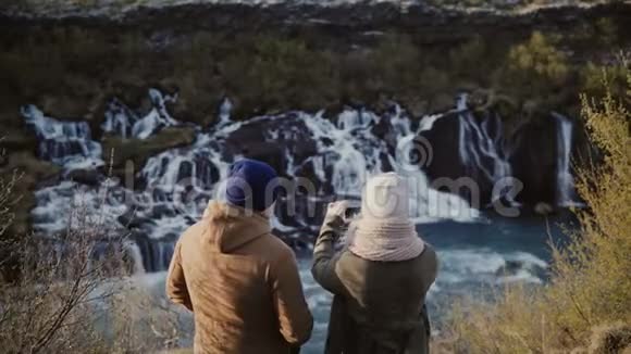 年轻的旅行夫妇站在冰岛瀑布附近的山上在智能手机上拍照视频的预览图