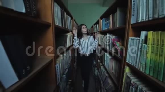漂亮的小女孩留着长长的黑发在图书馆的书架之间慢动作视频的预览图