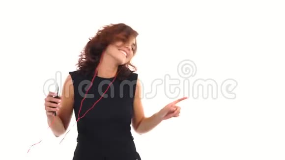 快乐的年轻女孩穿着黑色连衣裙用耳机听音乐在白色上独自跳舞玩得很开心视频的预览图