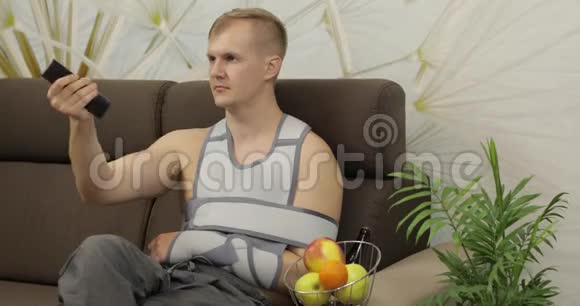 一个痛苦的男人胳膊断了戴着手臂支撑坐在沙发上看电视视频的预览图