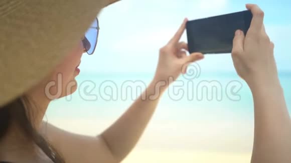 戴帽子的年轻旅行者女士在海边的智能手机拍照视频的预览图