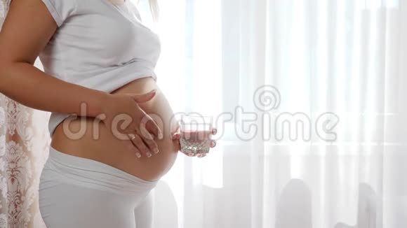 幸运的大肚子女性在白色的背景上拿着玻璃纯净水在裸露的腹部摩擦视频的预览图