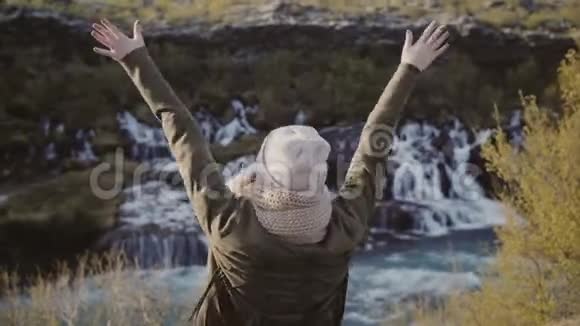 自由年轻女子站在冰岛瀑布附近举起双手摘下帽子感到快乐视频的预览图