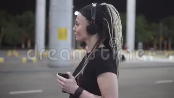 年轻有魅力的女人戴着恐惧症用智能手机在耳机上听音乐晚上在街上视频的预览图
