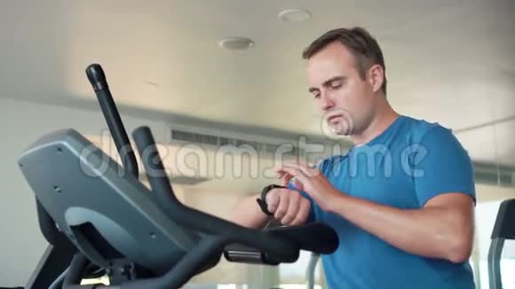 一个英俊健康的年轻人在健身房骑着健身车做运动用他的智能手表检查生命体征视频的预览图