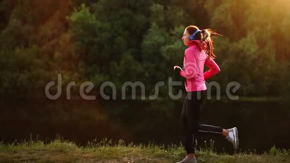 那个女孩在公园的日落时分沿着池塘跑步用耳机听音乐视频的预览图