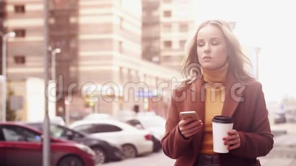 时髦的女人走在街上喝咖啡和使用智能手机视频的预览图