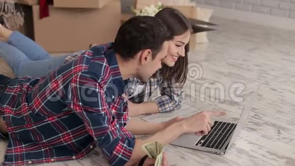 一对穿格子衬衫的幸福夫妇躺在一间新公寓的地板上手提电脑视频的预览图