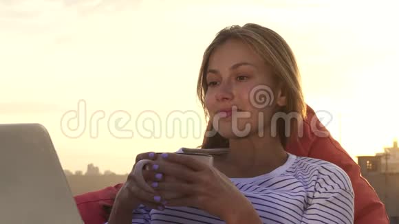 一个休息的女孩喝着温茶看着坐在屋顶上的笔记本电脑视频的预览图
