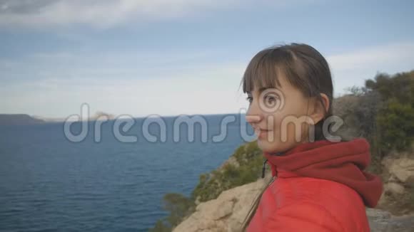 海上背景的年轻女子戴着红色连帽衫和泡泡夹克的白种人棕色头发的微笑女性视频的预览图