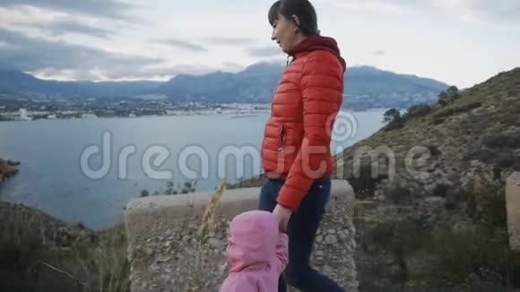迷人的年轻女子穿着红色泡泡夹克带着蹒跚学步的孩子走在海岸的山路上头戴玫瑰帽视频的预览图