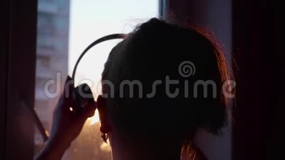 快关门一个年轻的女孩来到窗口戴上耳机听音乐背景模糊日落视频的预览图