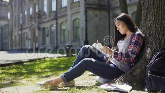 亚洲女孩坐在树下在笔记本上勾画周围环境艺术家视频的预览图
