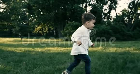 可爱的小男孩穿着白衬衫在草地上奔跑动作缓慢4k红色史诗日落视频的预览图