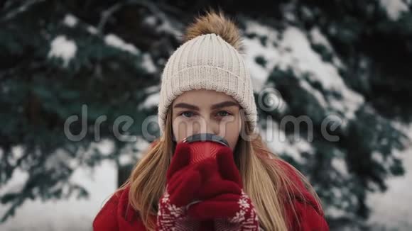 年轻漂亮的女孩穿着暖和的衣服站在雪中的圣诞树旁边从纸杯里喝咖啡视频的预览图