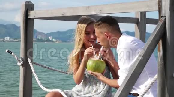 年轻的幸福夫妇坐在木墩上笑着喝着蓝色海洋背景上的椰子浪漫浪漫视频的预览图