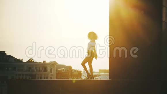 一位卷发的年轻女子在建筑物背景上表演迷人的脱衣舞日落视频的预览图