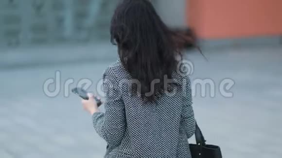 年轻的商务小姐穿着一件紧身夹克头发深棕色手里拿着智能手机走在城市的大街上视频的预览图