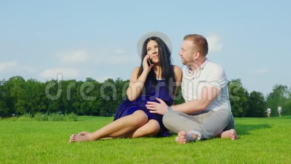 年轻的孕妇通过电话说话她旁边坐着她的丈夫一起在公园里休息视频的预览图