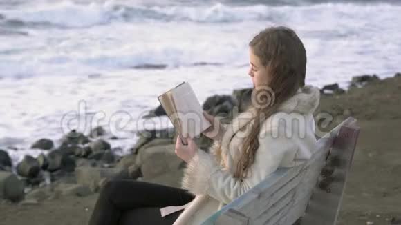 一个年轻的女孩坐在海边的长凳上在傍晚的日落时读一本书4K一个孩子在海滩上看书视频的预览图