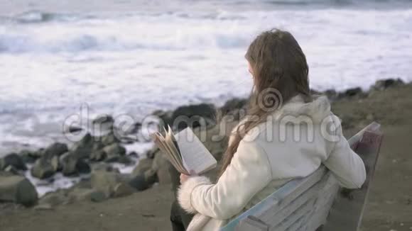 一个年轻的女孩坐在海边的长凳上在傍晚的日落时读一本书4K一个孩子在海滩上看书视频的预览图