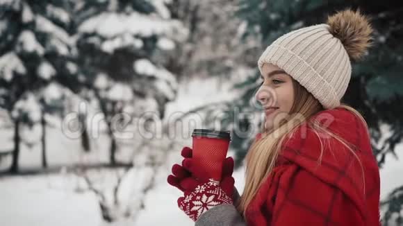 年轻漂亮的女孩穿着暖和的衣服站在圣诞树旁边在雪地里喝着报纸上的热饮料视频的预览图