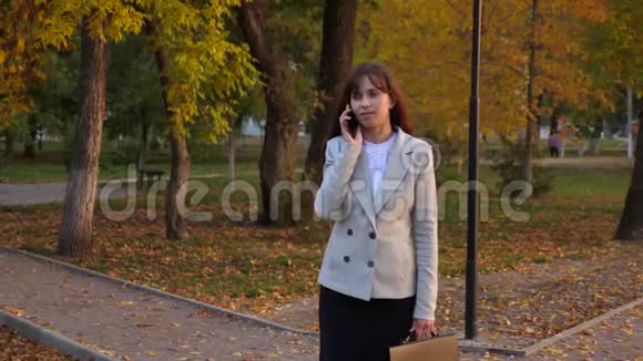 美丽的女人用手机说话快乐的女孩走过秋天的公园微笑着视频的预览图