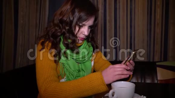 年轻漂亮的女孩坐在餐桌旁的咖啡馆里用智能手机她微笑着一条绿色围巾视频的预览图