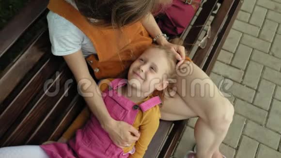 妈妈带着女儿学龄前女孩躺在公园里一个年轻女人的腿上的长凳上夏日晴天视频的预览图