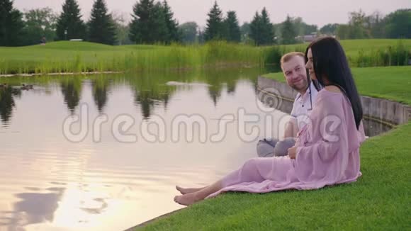 与丈夫穿粉色连衣裙的孕妇他们坐在湖边的草地上视频的预览图