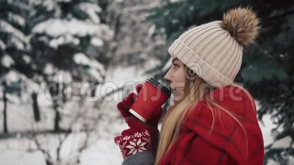 年轻漂亮的女孩穿着暖和的衣服站在圣诞树旁边在雪地里喝着报纸上的热饮料视频的预览图