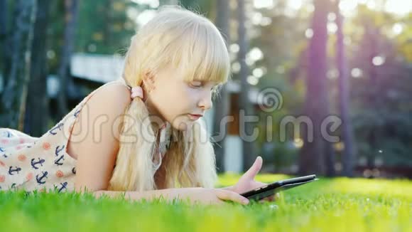 金发女孩6年在平板电脑上玩情绪反应躺在房子后院的草坪上侧视图视频的预览图
