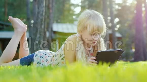 金发女孩6年在平板电脑上玩情绪反应躺在房子后院的草坪上侧视图视频的预览图