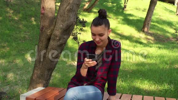 年轻漂亮的女孩穿着格子衬衫坐在公园的长凳上在网上聊天中积极改写视频的预览图